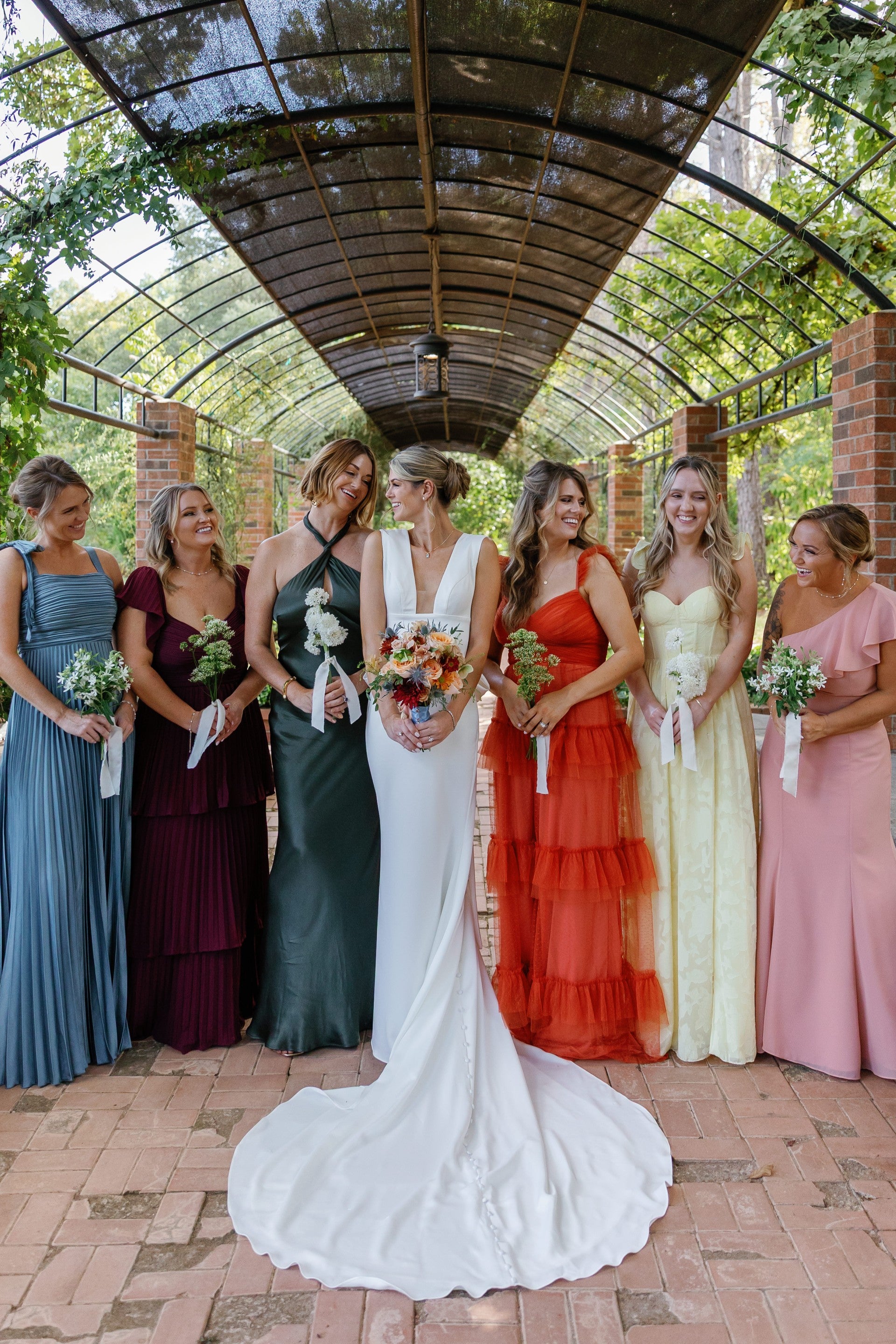 Bride in a white dress with bridesmaids in colorful dresses under a glass roof.
